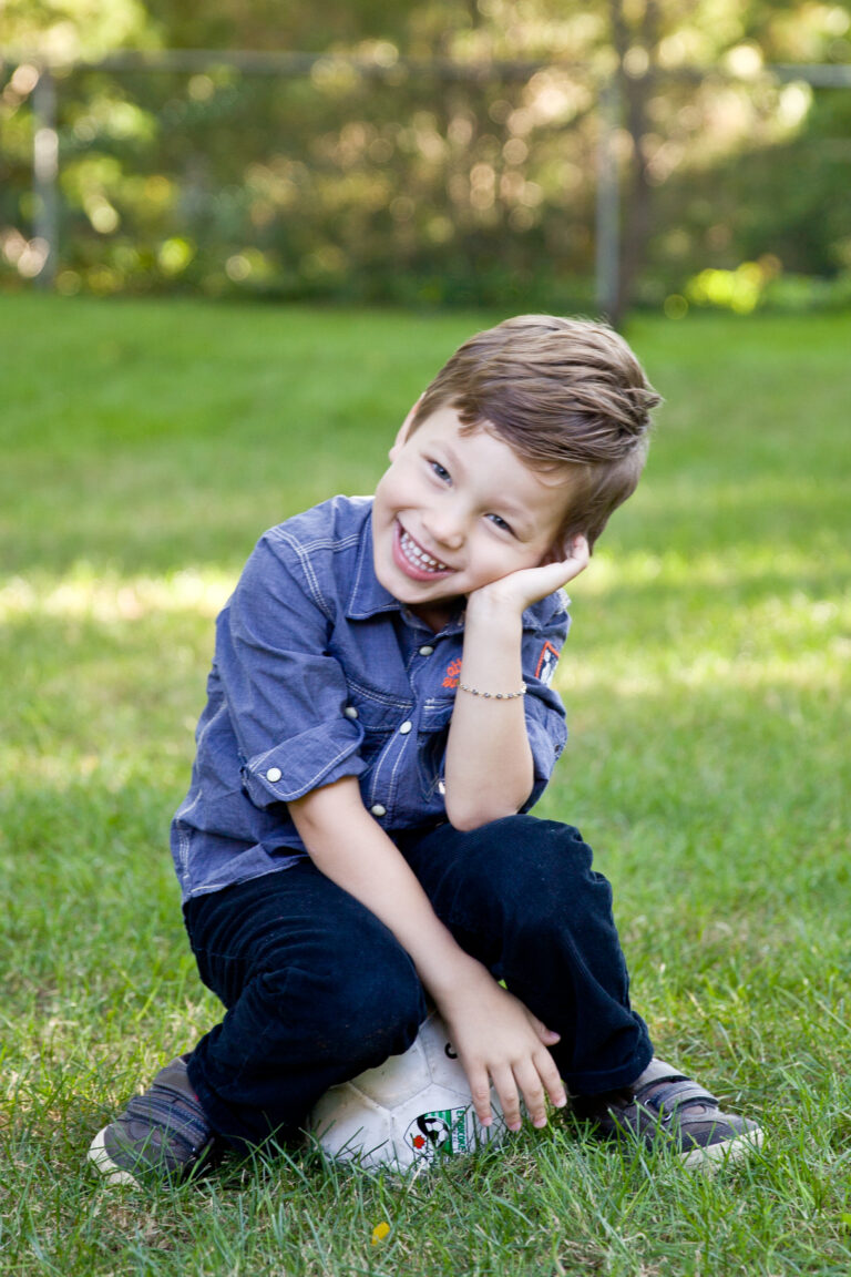 young boy sitting on a soccer ball in the grass posing for a photo, while laughing